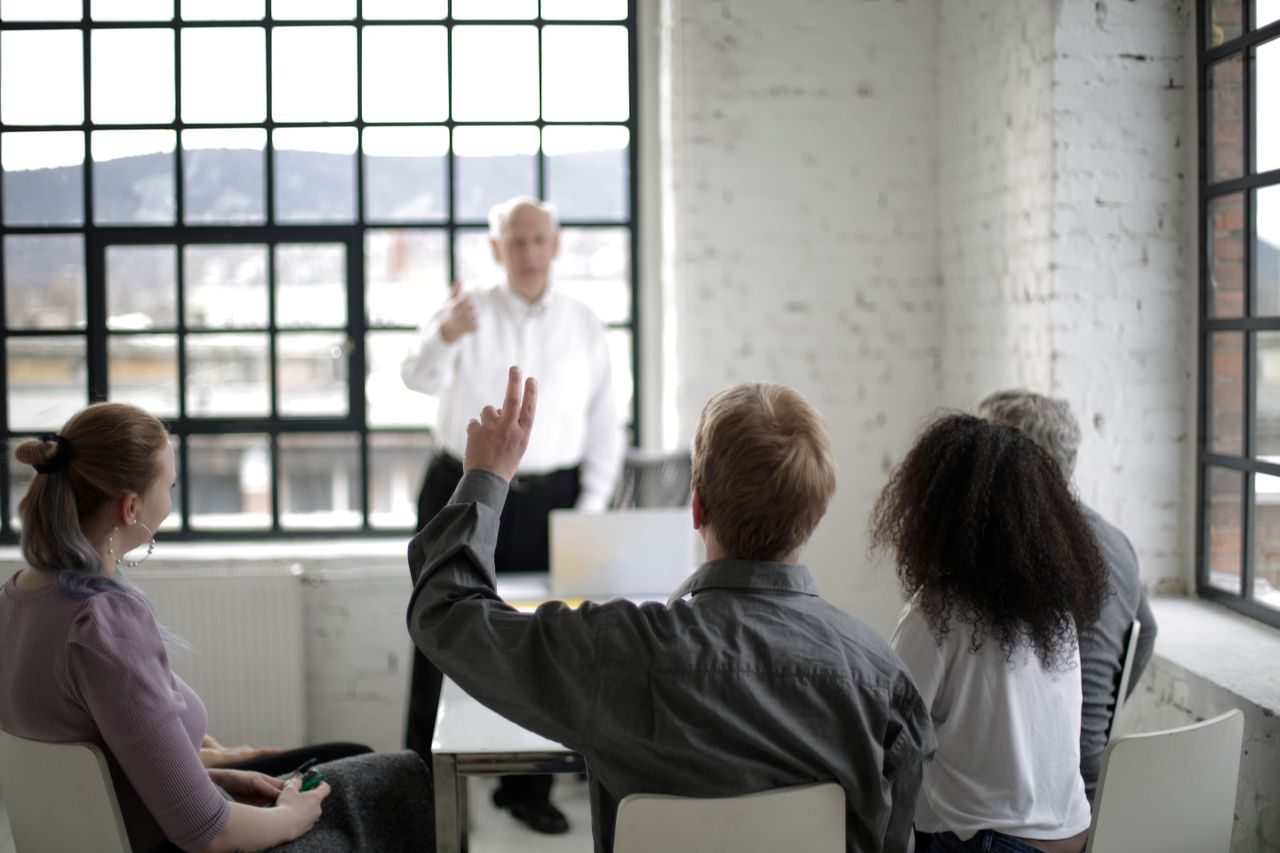 who-we-are Group of adults in a discussion, with one person raising a hand in a bright office space.